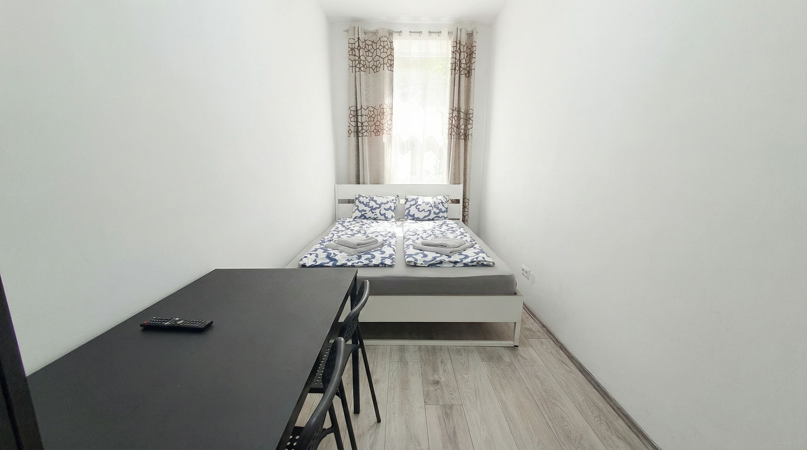 A small, narrow room with a bed at the end, a desk with a chair, and a window with curtains.