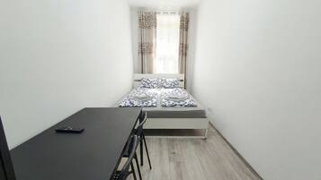 A small, narrow room with a bed at the end, a desk with a chair, and a window with curtains.