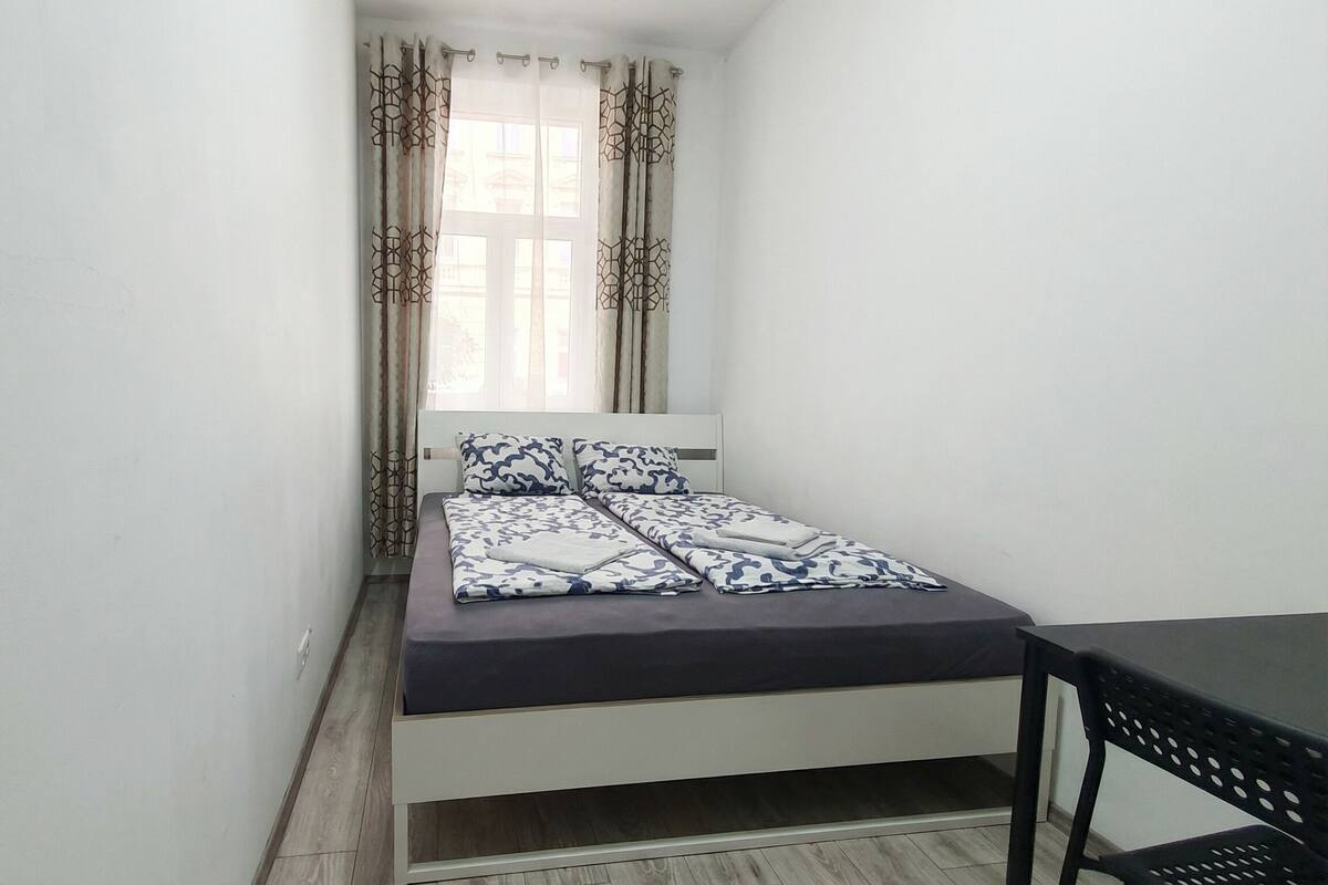 Una habitación pequeña con cama individual, piso de madera, cabecera blanca y ropa de cama estampada.