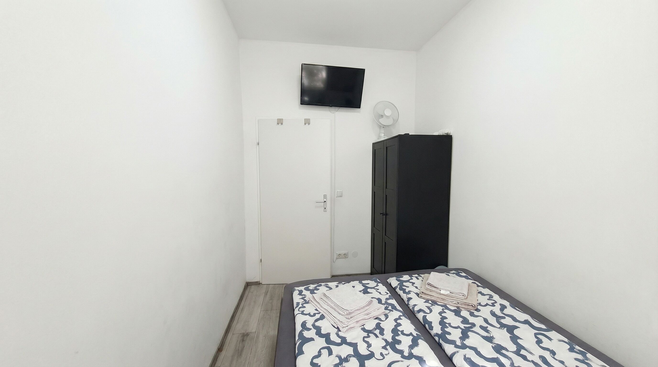 A small, narrow room with a bed, a television mounted on the wall, and a wardrobe.