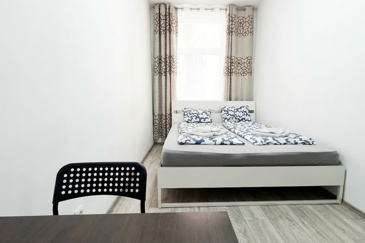 Una habitación pequeña con una cama individual, cabecera blanca, una silla negra y una ventana con cortinas estampadas.