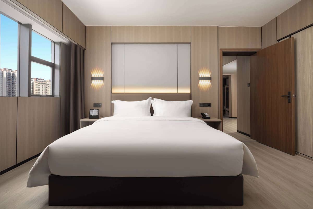 A modern hotel bedroom with a large bed, bedside lamps, a nightstand, and a view of the city through large windows.