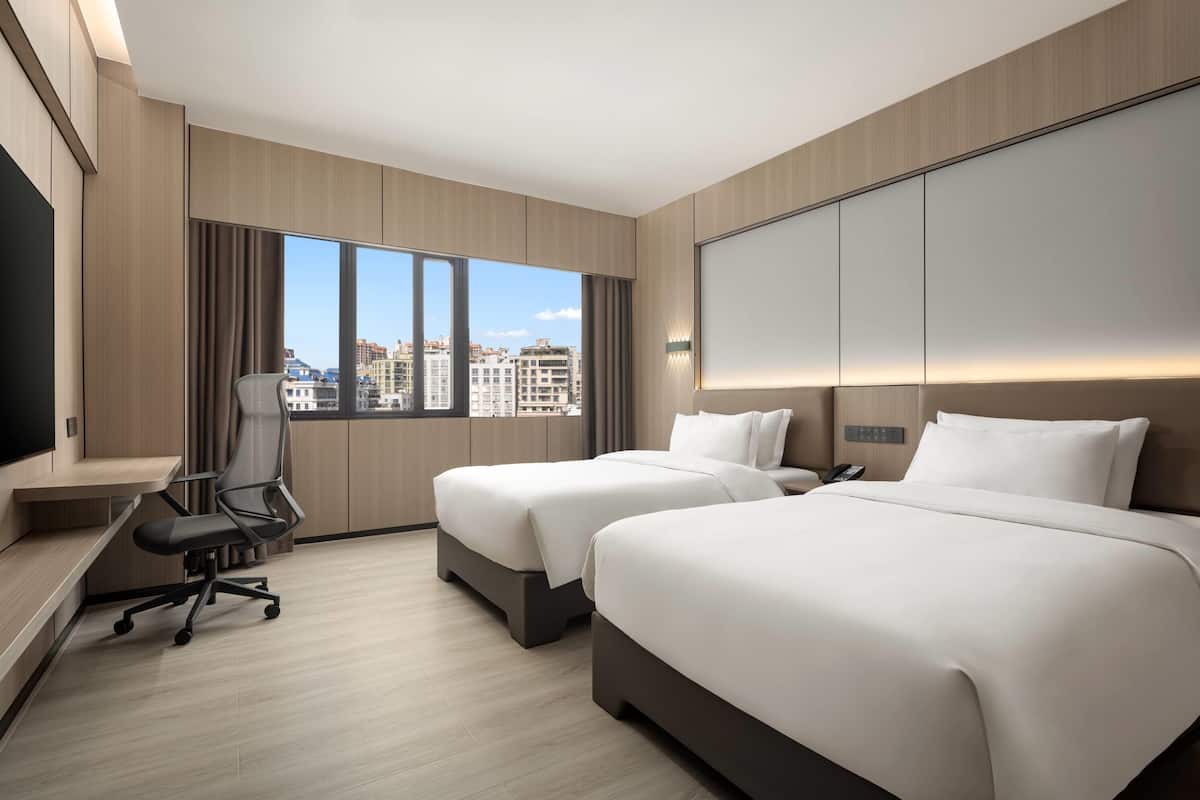 A hotel room with two beds, a desk, a chair, and a large window with a city view.