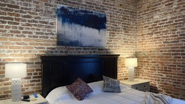 Een slaapkamer met een bakstenen muur, een donker hoofdeinde, een wit sprei, twee bedlampen en een blauw abstract schilderij.