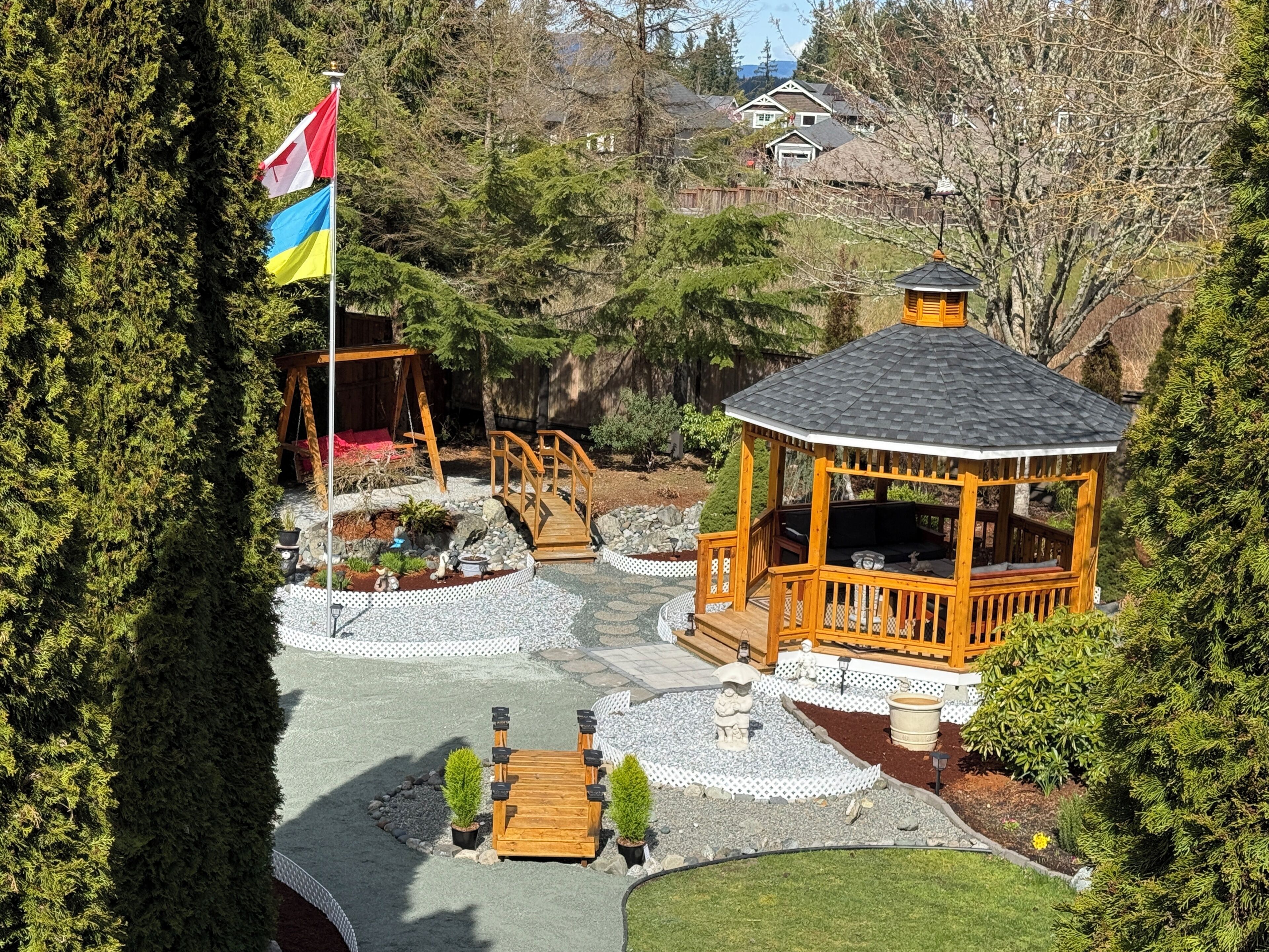 Ein Pavillon mit Holzterrasse, umgeben von einem Garten mit einer kleinen Brücke und einer Fahnenstange.
