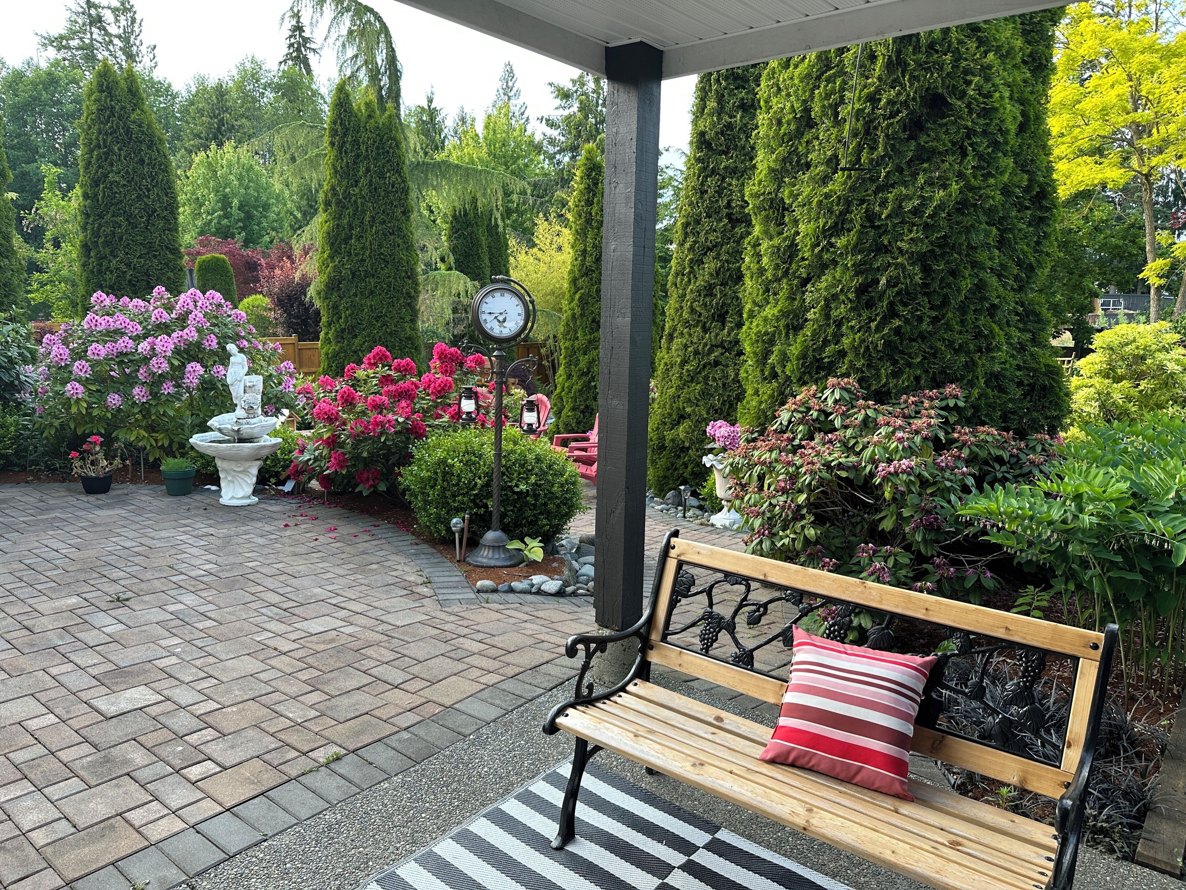 Een patio met een houten bankje, een klok en verschillende potten met planten en bloemen.