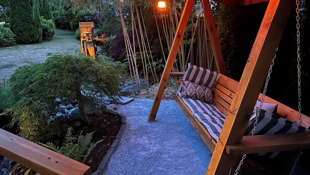 A wooden swing with cushions, surrounded by a garden with a gravel path, and a wooden deck. - Charming Mill Bay 2 Br Suite (Mill Bay)