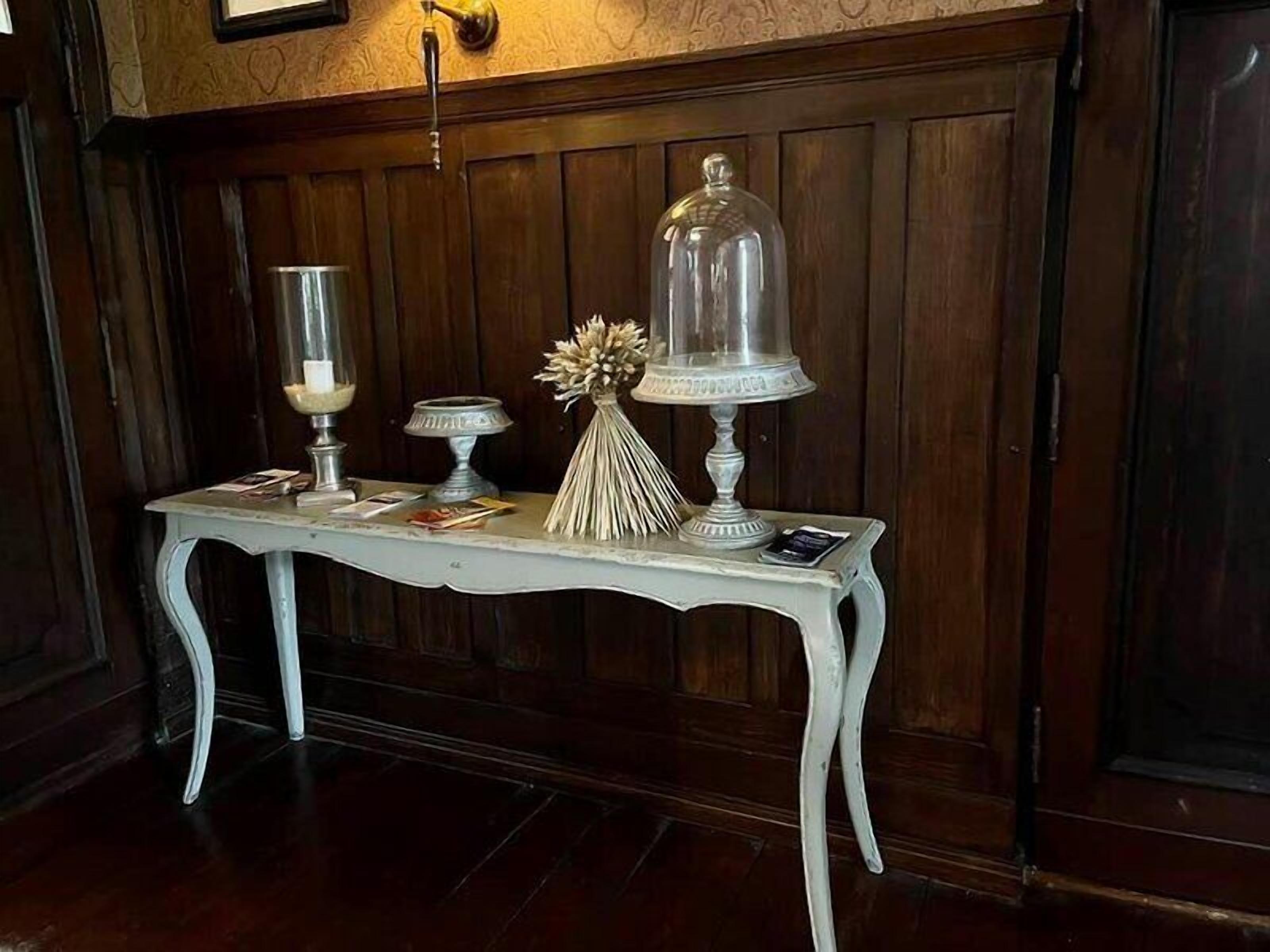 A white table with a glass dome centerpiece, a candle holder, and a decorative vase against a wooden wall.