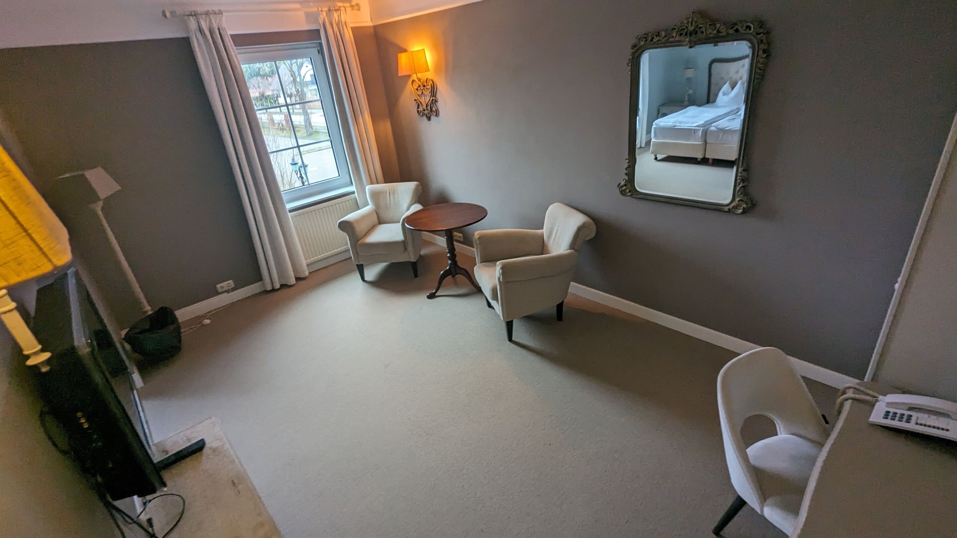 A hotel room with a bed, two armchairs, a small round table, a lamp, a mirror, and a window with curtains.