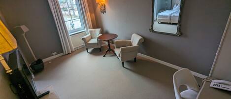Ett hotellrum med en säng, två fåtöljer, ett litet runt bord, en lampa, en spegel och ett fönster med gardiner.