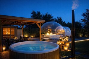 Un jacuzzi y un dormitorio con paredes de vidrio y chimenea, rodeado de iluminación exterior y una terraza de madera.