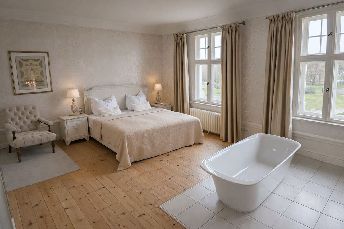 Ein Schlafzimmer mit einem großen Bett, einem Sessel, einem Nachttisch, einer Badewanne und einem Fenster mit Vorhängen.