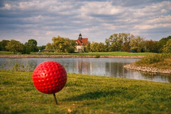 Una pelota de golf en primer plano, con un lago y una casa al fondo.