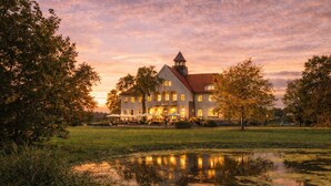 Ein großes, historisches Gebäude mit einem markanten Turm, umgeben von einem Teich und Bäumen, die den Sonnenuntergang reflektieren.