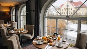 Ein Essbereich mit runden Tischen, Stühlen und Frühstücksangeboten. Große Fenster bieten Blick auf ein Gebäude mit rotem Dach.