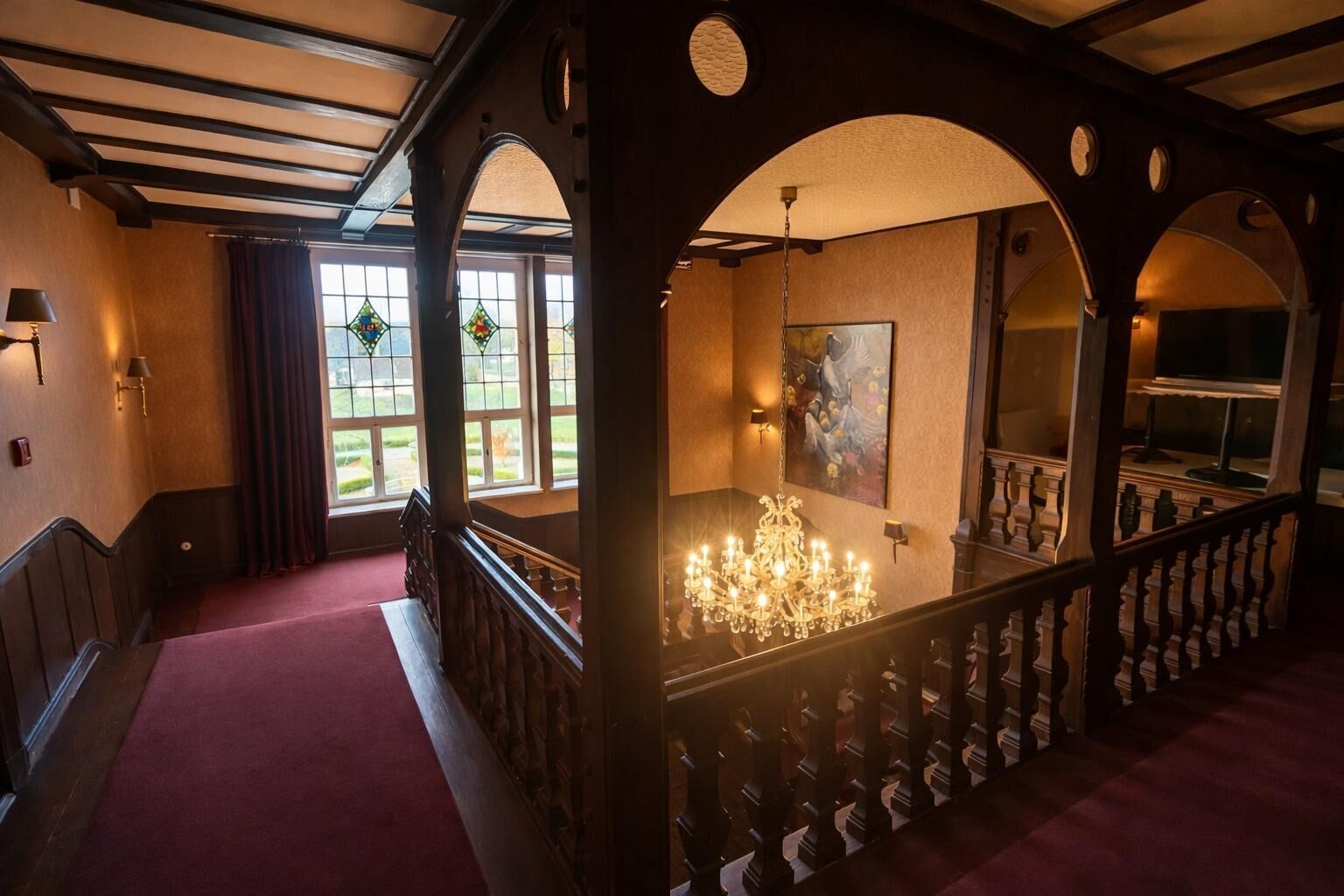 A grand staircase with a chandelier, arched wooden railings, and a view of a garden through large windows.