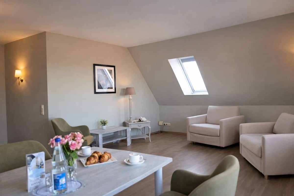 Ein modernes, helles Wohnzimmer mit Essbereich, ein Tisch gedeckt mit Blumen, Gebäck und einer Tasse Kaffee. Zwei Sofas, eine Lampe und ein gerahmtes Bild an der Wand.