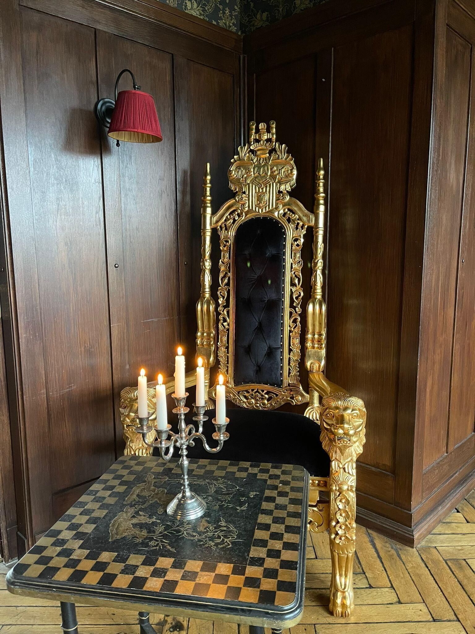 Ein Zimmer mit einem karierten Tisch, einem goldenen, verzierten Sessel, einem Kandelaber mit brennenden Kerzen und einer roten Wandleuchte.