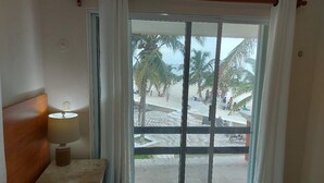 A room with a large sliding glass door leading to a beach view, a bedside table with a lamp, and a wooden headboard. - Casa Playasol (Puerto Morelos)