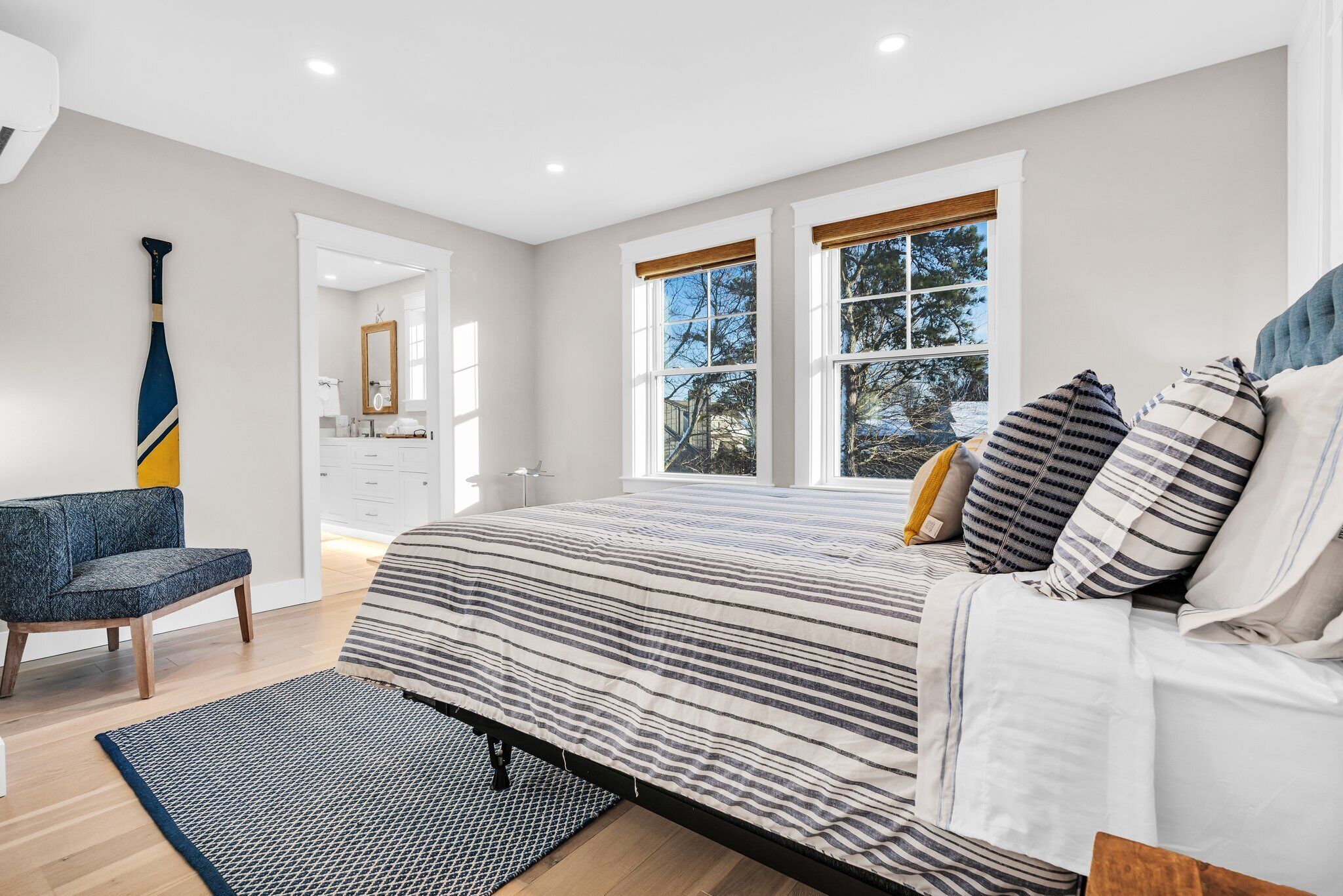 A modern bedroom with a large bed, a chair, and a view of the outdoors through two windows.