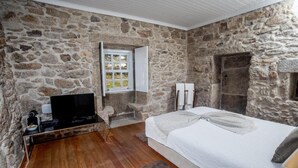 Une chambre avec des murs en pierre, un plancher en bois, un lit recouvert d’une courtepointe blanche, un téléviseur posé sur un meuble et une fenêtre avec des rideaux.