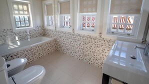 Une salle de bain équipée d’une baignoire, de toilettes et d’un lavabo, avec un papier peint à motifs et plusieurs fenêtres dotées de stores.