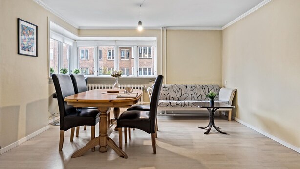 Un coin repas comprenant une table ronde en bois, quatre chaises noires, une petite table d’appoint et un canapé agrémenté d’un coussin à motifs.