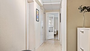 Un couloir étroit avec une porte donnant sur une pièce. On y trouve un tableau encadré accroché au mur, un petit banc et une commode avec des tiroirs.