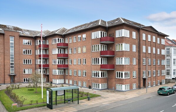 Un immeuble résidentiel à plusieurs étages, doté d’une façade en briques, de nombreux balcons et d’une pelouse verdoyante agrémentée d’un banc.