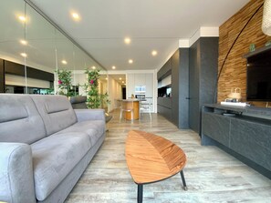 Una sala moderna con un sofá gris, una mesa de centro de madera y una zona de cocina con televisor y paneles de pared de madera.