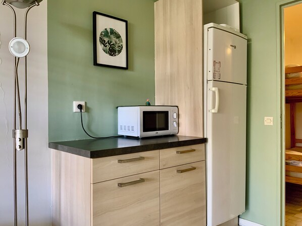 A kitchenette with a microwave, a refrigerator, a wooden cabinet, and a framed picture on the wall. - Le Santa Maria - Appartement avec jardin privé & parking | Port Camargue (Le Grau-du-Roi)