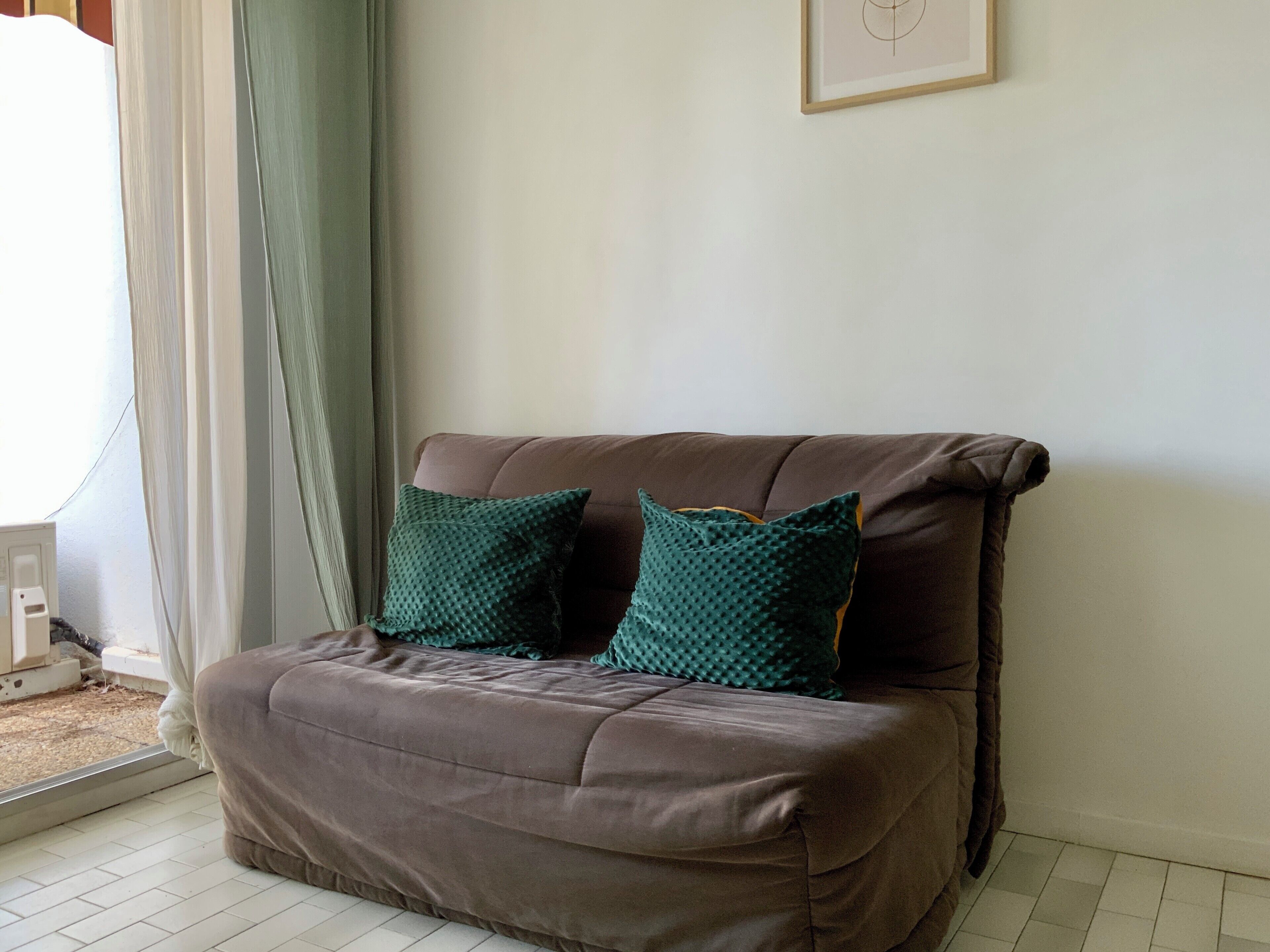 Un sofá marrón con dos almohadones verde azulado en una habitación con pared blanca y un cuadro enmarcado.