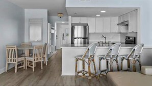 A modern kitchen with white cabinets, a stainless steel refrigerator, and a central island with bar stools. - Stunning Beachfront Condo with Pool (Surf City)
