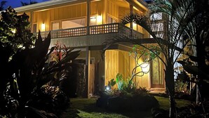 A two-story house with a balcony, surrounded by tropical plants and a well-lit entrance. - Tropical Ocean View! Escape to the Rainforest getaway of your dreams! (Keaau)