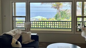 A room with a large window overlooking a coastal view, a dark blue sofa with a white throw blanket, and a white table with a lamp. - Tropical Ocean View! Escape to the Rainforest getaway of your dreams! (Keaau)