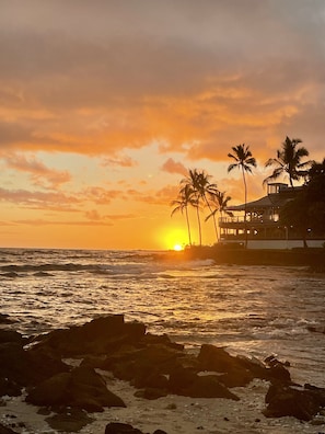 A beachfront property with a view of the ocean at sunset, featuring palm trees and a building with a balcony.