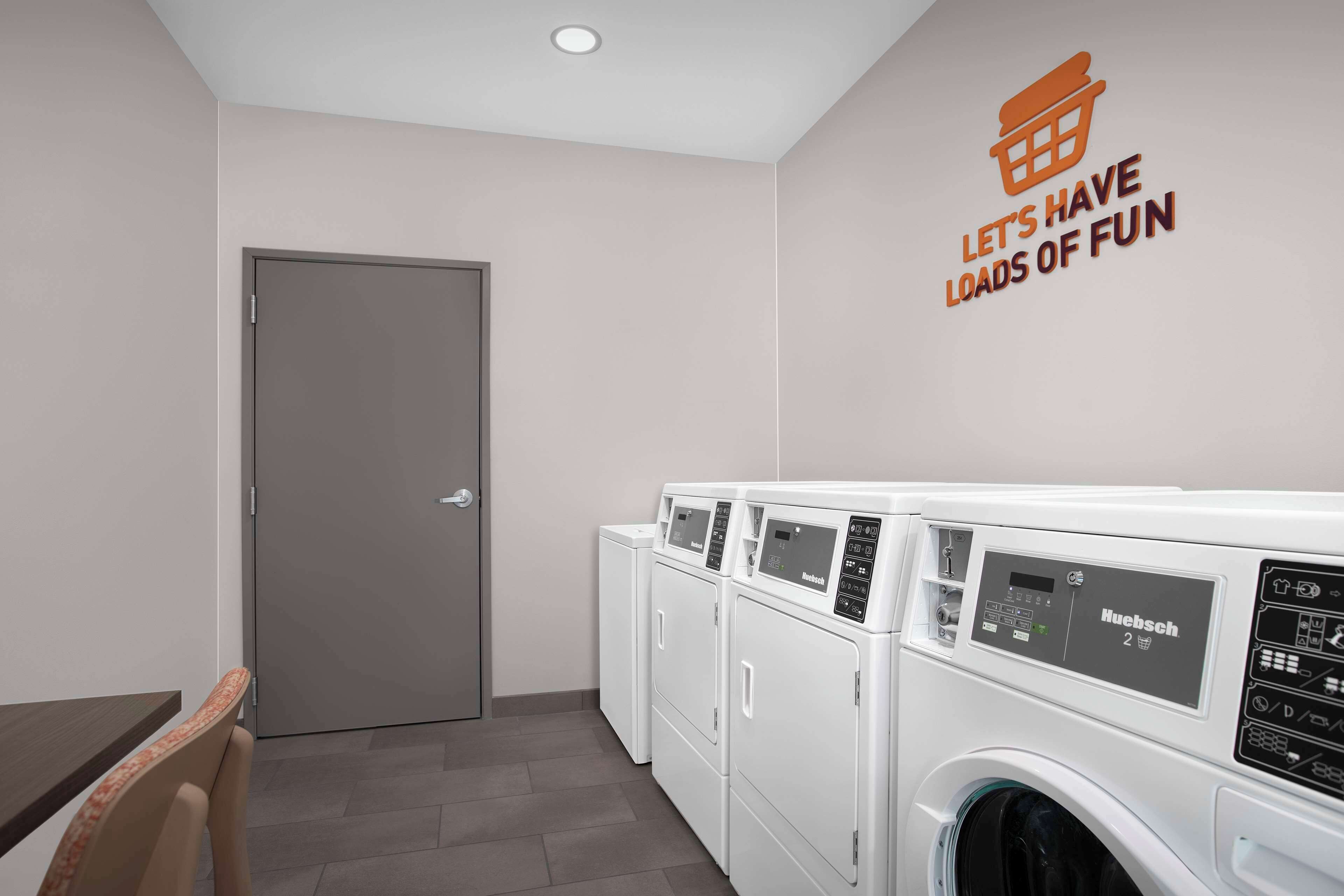 A laundry room with a row of white Nuensch washing machines, a door, and a wall sign that reads "LET'S HAVE LOADS OF FUN".