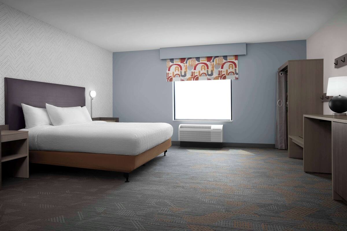 A hotel room with a bed, bedside table, lamp, wardrobe, and a window with a patterned curtain.
