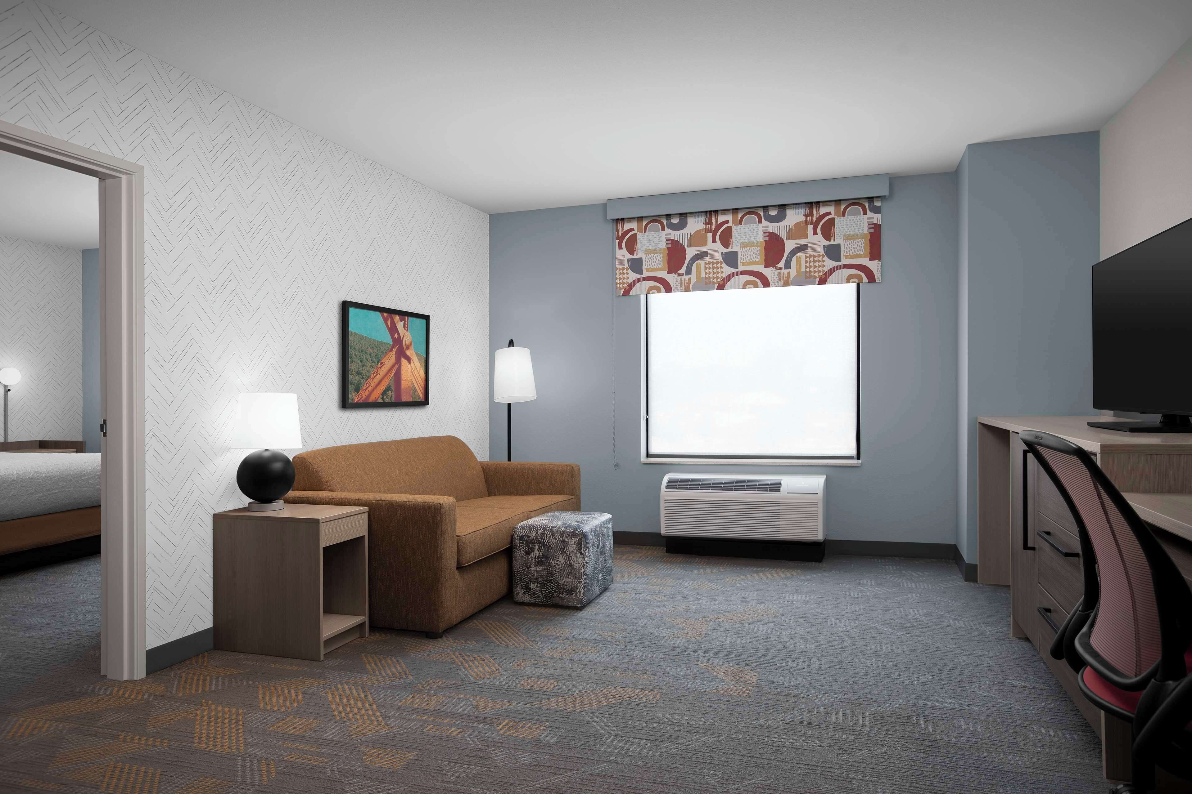 Ein Hotelzimmer mit Bett, Sofa, Schreibtisch mit Lampe, Fernseher und Fenster mit gemustertem Vorhang.