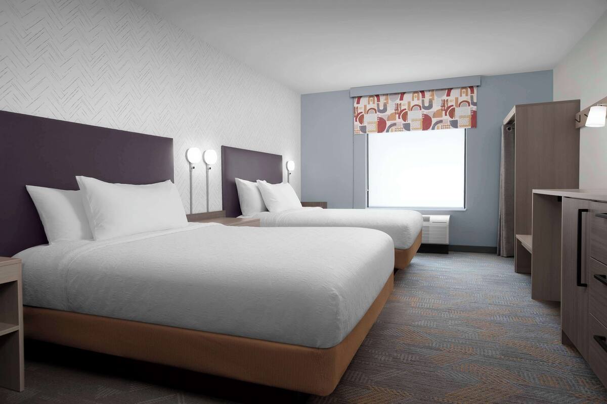 A hotel room with two beds, a desk, a wardrobe, and a window with a patterned curtain.