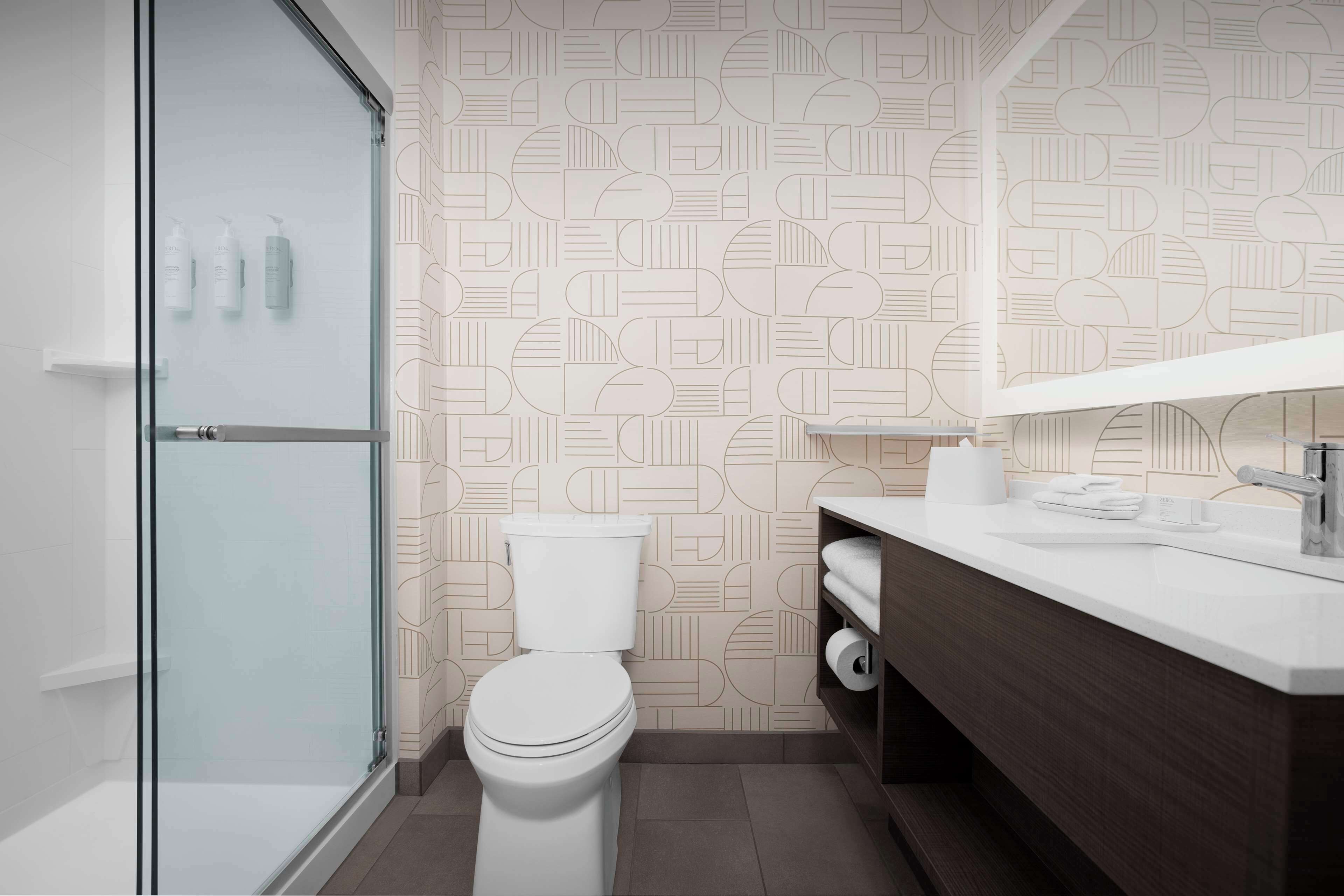 Un baño moderno con pared de diseño geométrico, cabina de ducha de cristal, inodoro blanco, tocador de madera con espejo y lavabo blanco.