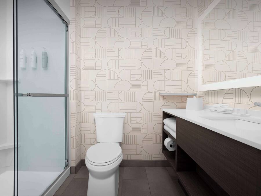 Un baño moderno con pared de diseño geométrico, cabina de ducha de cristal, inodoro blanco, tocador de madera con espejo y lavabo blanco.
