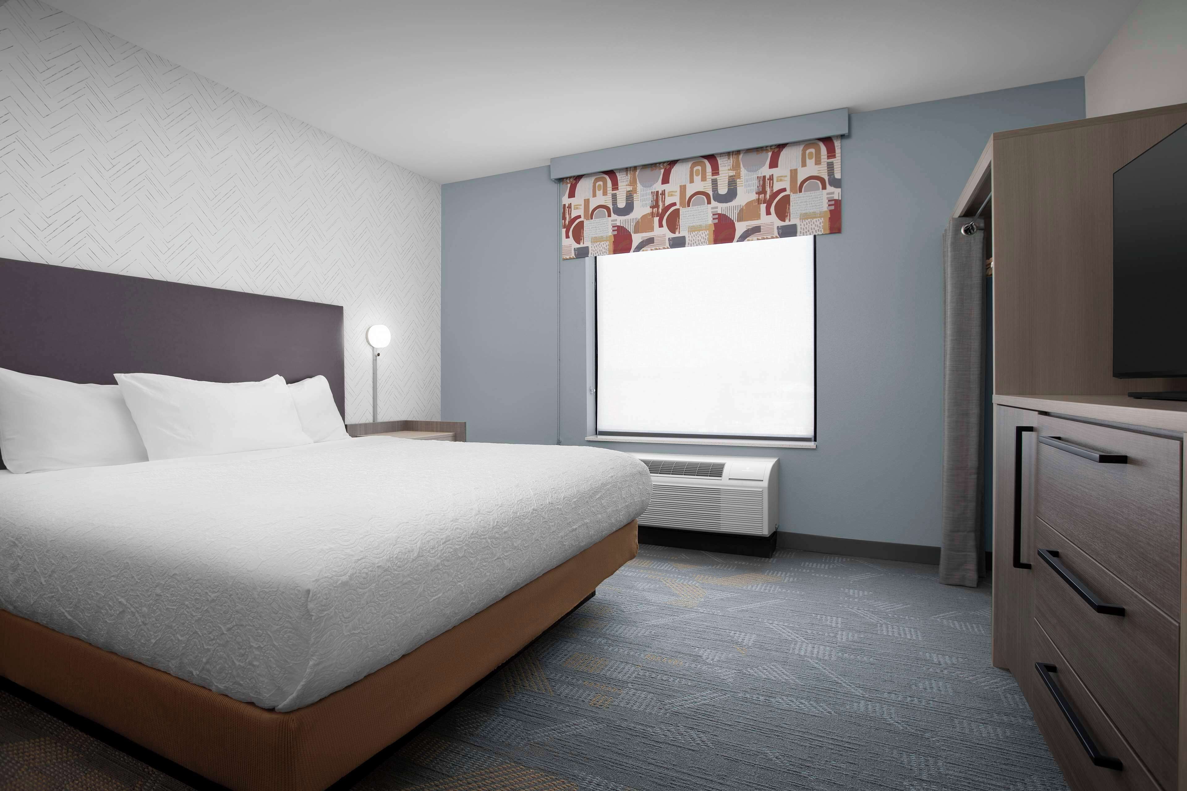 Quarto de hotel com cama, escrivaninha, televisão e janela com cortina.