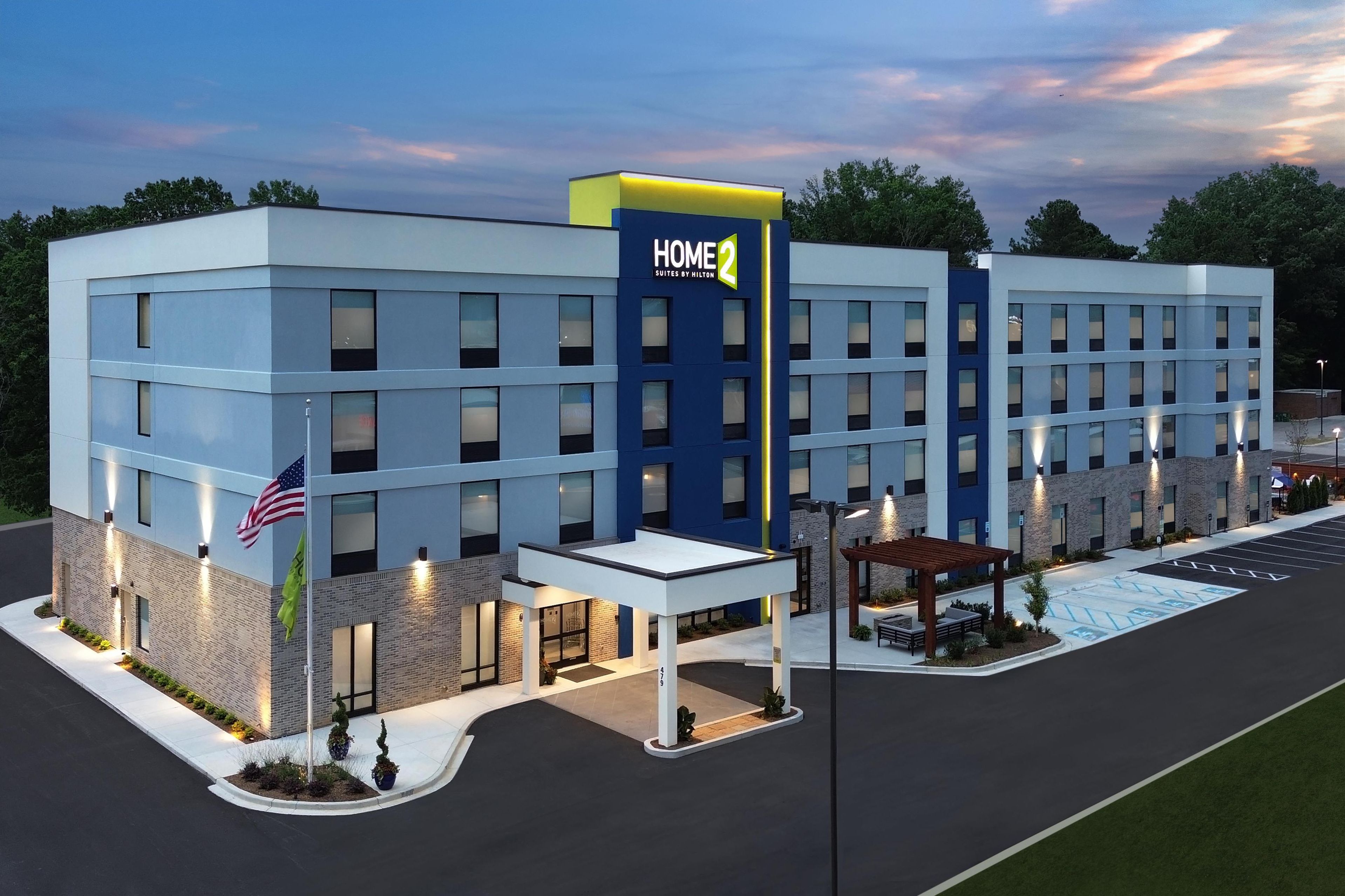 Un hotel moderno con un llamativo letrero 'HOME 2', múltiples accesos y una zona de estacionamiento bien iluminada.