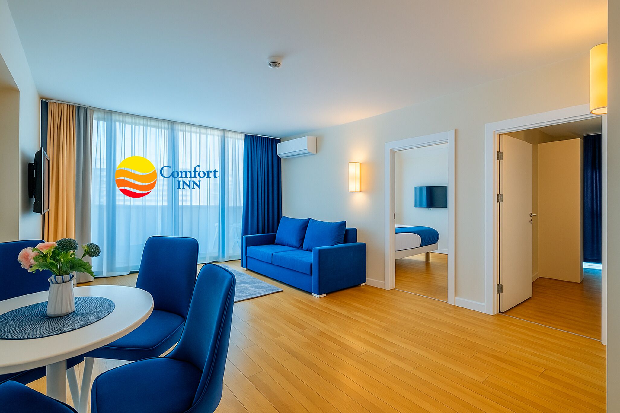 A modern hotel room with a blue sofa, a round table with a vase of flowers, and a large window with curtains.