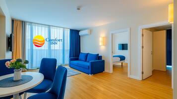 A modern hotel room with a blue sofa, a round table with a vase of flowers, and a large window with curtains.