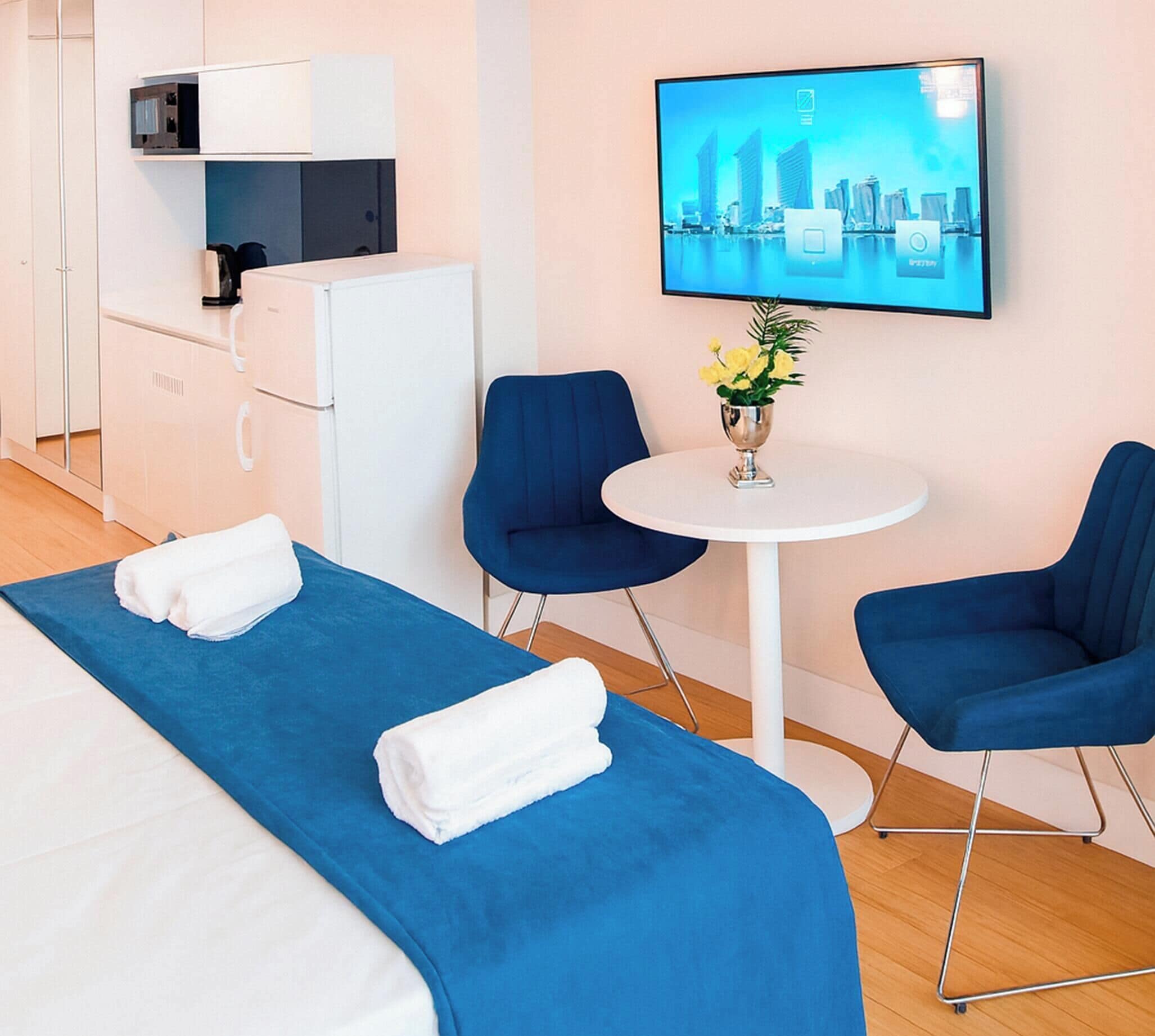 Une chambre d’hôtel avec un lit, deux chaises, une petite table avec un vase et un téléviseur diffusant des images de la ville.