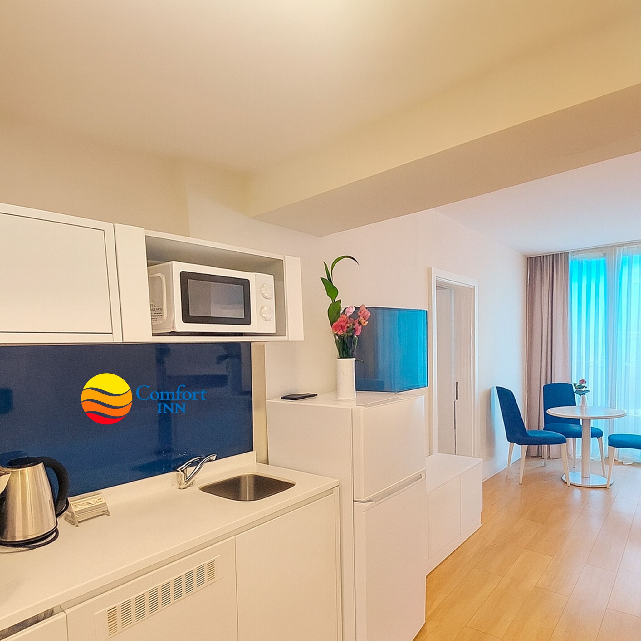 A modern kitchen with a microwave, sink, and a logo for Comfort Inn. There is a dining area with blue chairs and a table.