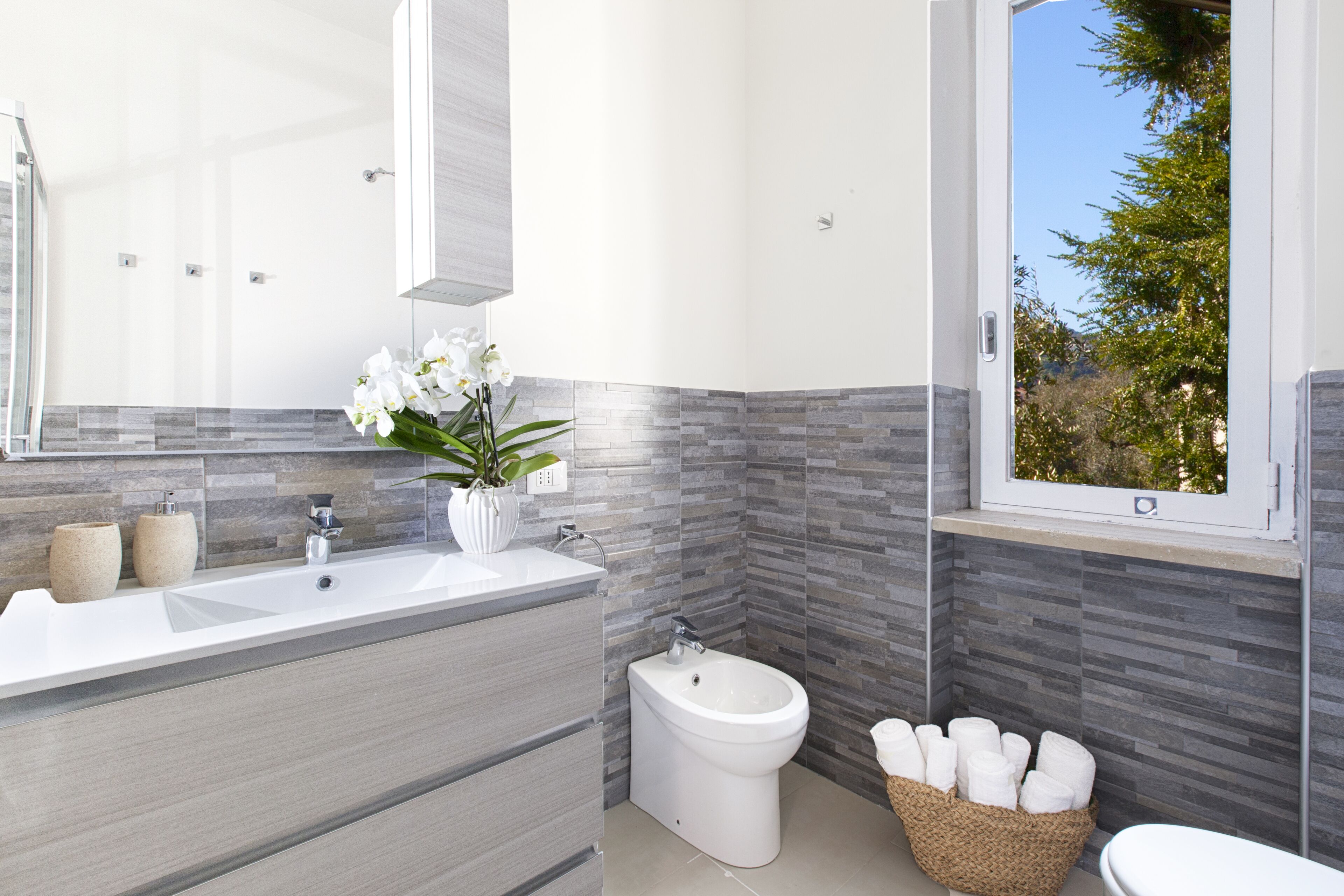 A modern bathroom with a large mirror, a bidet, a sink, and a window with a view of trees.