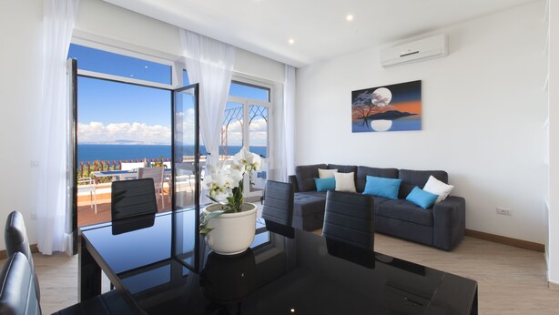 A modern living room with a glass dining table, black chairs, a dark blue sofa with blue and white cushions, and a large window offering a view of the sea and a bridge.